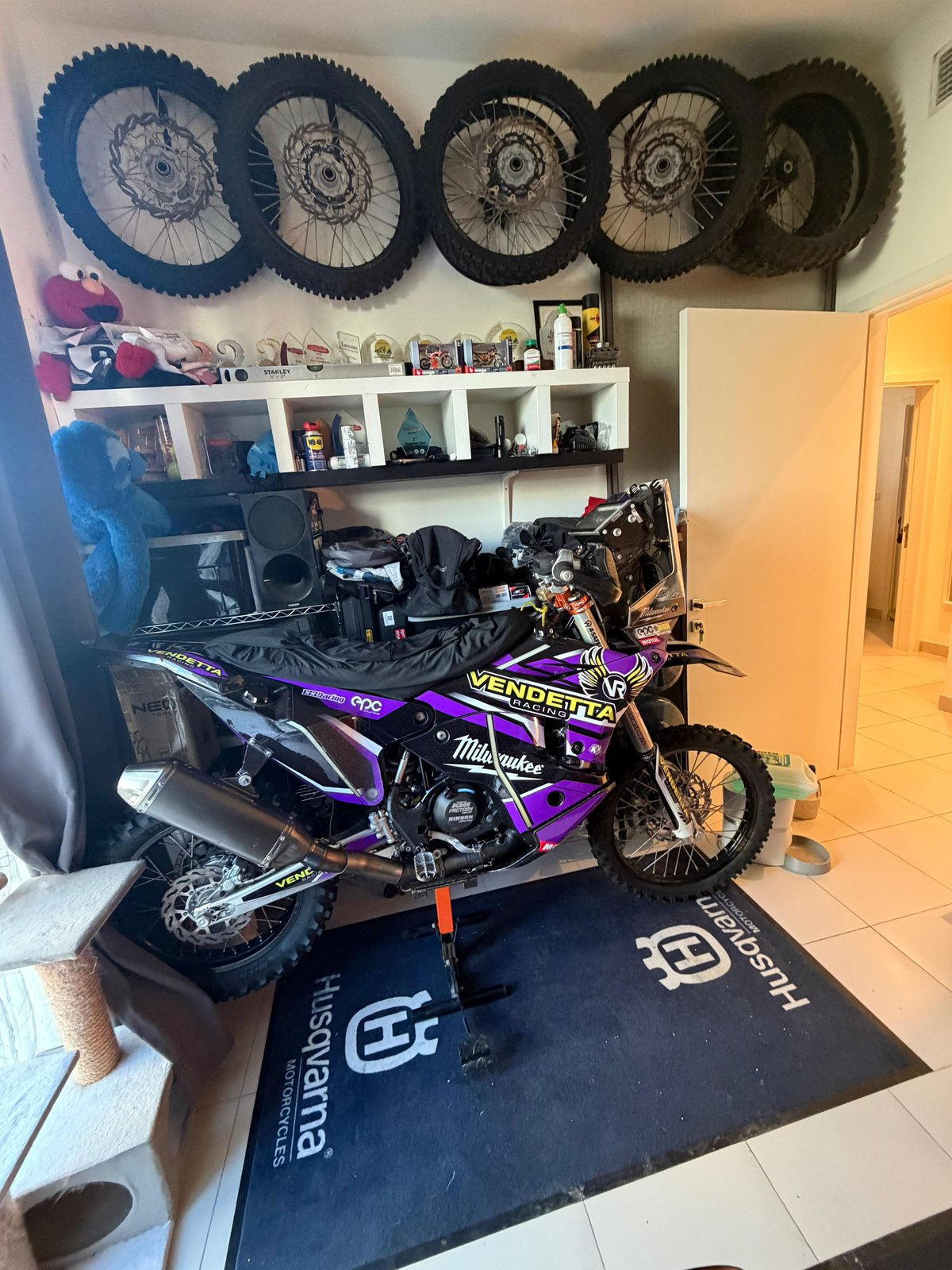 Home garage with dirt-bike wheels mounted on the wall, trophies and Cookie Monster on the shelf, race bike on a Husqvarna mat