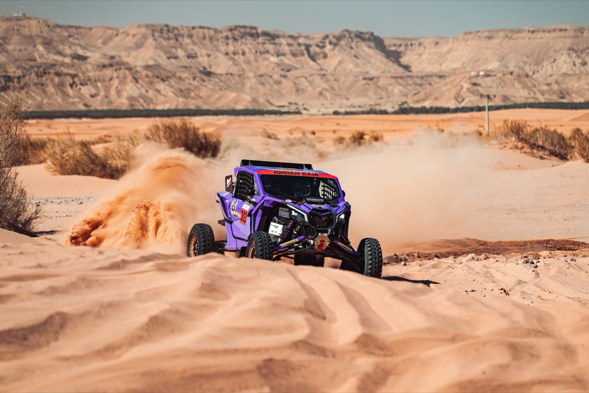 Vendetta Racing UTV #303 cresting a desert dune with rocky escarpments behind it.