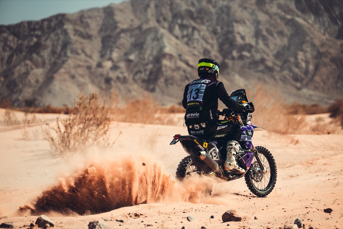 Rally raid motorcycle #13 in Vendetta livery roosting sand against a Wadi Rum mountain backdrop.